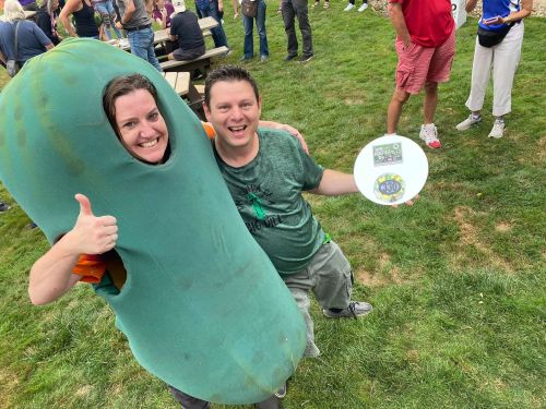 A person in a pickle costume gives a thumbs up next to a smiling man holding a frisbee at an outdoor event.