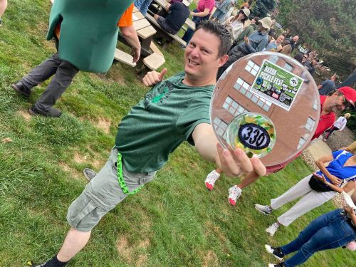 A man in a green shirt proudly holds a circular award at an outdoor event, surrounded by a crowd.