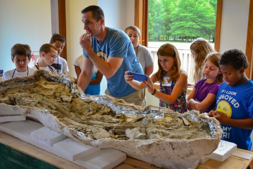 A group of children observes a man explaining a large fossil display in a classroom setting.