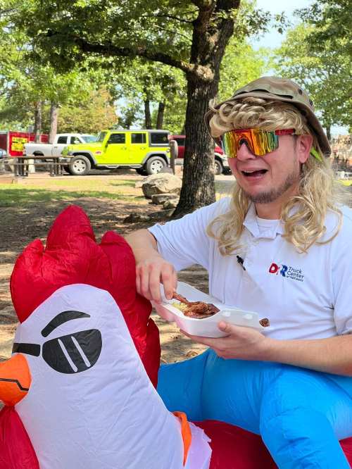 A person with a mullet wig and sunglasses sits on an inflatable chicken, enjoying food outdoors.