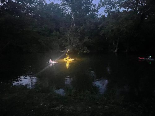 Two kayakers paddle on a dark river, illuminated by lights, surrounded by trees and a serene nighttime atmosphere.