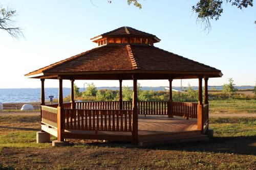 A wooden gazebo with a shingled roof overlooking a calm lake and grassy area under a clear blue sky.