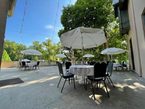 Outdoor dining area with white umbrellas and tables, surrounded by trees and string lights on a sunny day.