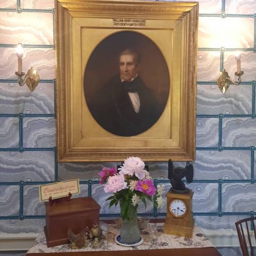 A vintage table with a flower vase, clock, and a box, beneath a portrait of William Henry Harrison on patterned wallpaper.
