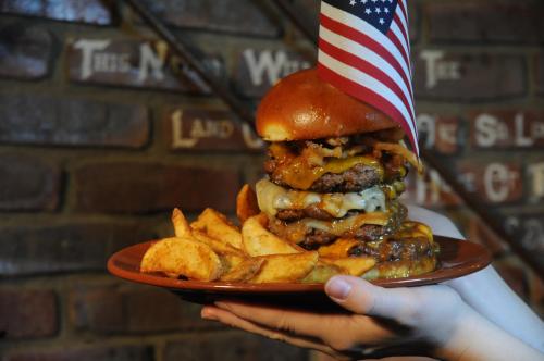 A towering burger with multiple patties, cheese, and crispy onions, served with potato wedges and an American flag.