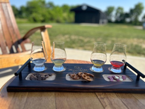 A tasting flight of four whiskey glasses on a wooden tray, accompanied by chocolate, pretzels, and candies outdoors.