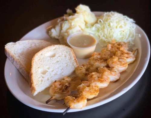 A plate with shrimp skewers, mashed potatoes, shredded cabbage, two slices of bread, and a small cup of sauce.