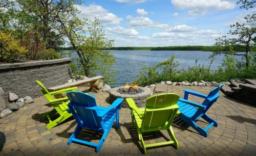 Colorful adirondack chairs surround a fire pit by a serene lake, with lush greenery and a blue sky in the background.