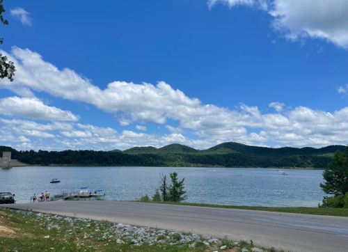 A serene lake surrounded by green hills under a blue sky with fluffy clouds, with a road and boats in the foreground.