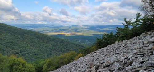 A panoramic view of rolling green hills and valleys under a partly cloudy sky, with a rocky foreground.