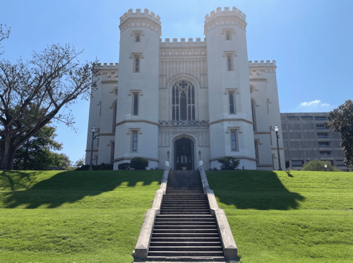 Historic castle-like building with towers, surrounded by green grass and trees, under a clear blue sky.