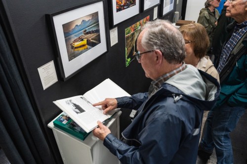 A man examines a book while standing near framed photographs in an art exhibit, with visitors in the background.
