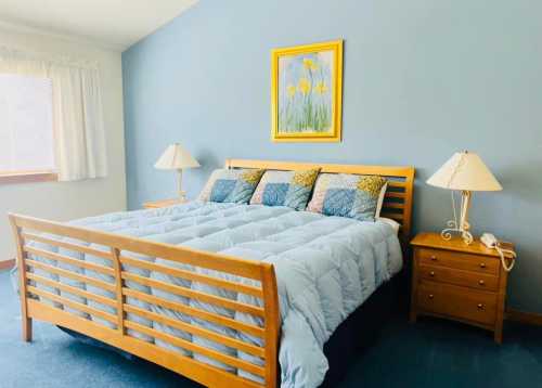 A cozy bedroom featuring a wooden bed with blue bedding, two lamps, and a floral painting on the wall.