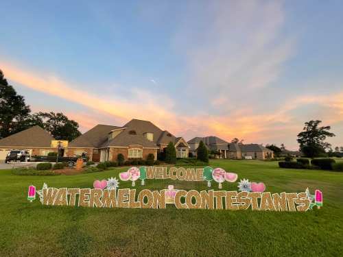 A colorful sign reading "Welcome Watermelon Contestants!" in front of houses at sunset, with a grassy lawn.