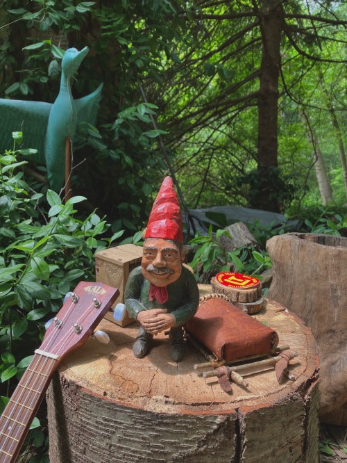 A whimsical garden gnome sits on a wooden stump surrounded by greenery, with a guitar and various rustic items nearby.
