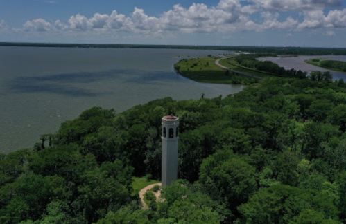 Aerial view of a tall tower surrounded by lush greenery near a lake and winding road under a cloudy sky.
