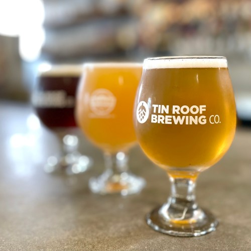 Three glasses of craft beer from Tin Roof Brewing Co. in a row, showcasing different colors and styles on a bar.