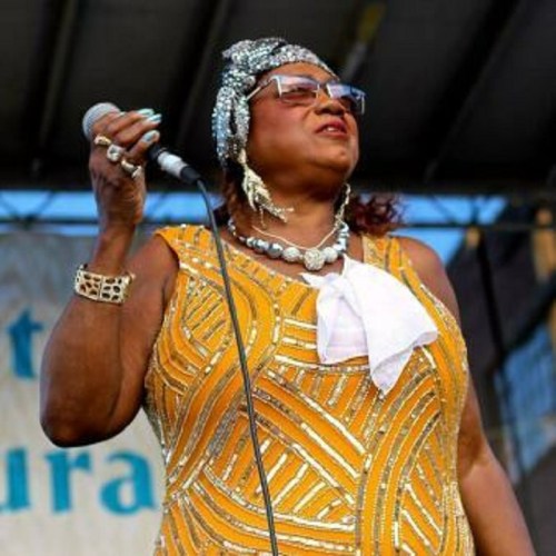 A performer in a sparkling yellow dress sings into a microphone, adorned with jewelry and sunglasses.