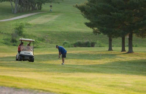 A golfer prepares to swing while another person sits in a golf cart on a lush green course.