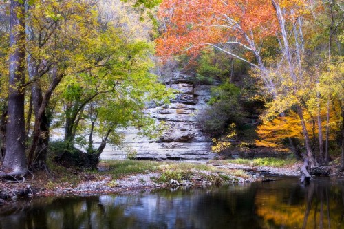 A serene river scene with colorful autumn trees and a rocky cliff reflecting in the water.