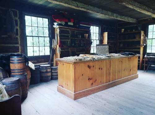 Interior of a rustic wooden room with a central table, shelves, barrels, and natural light from large windows.