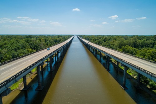 Aerial view of a long bridge over a wide waterway, surrounded by lush greenery and blue sky.