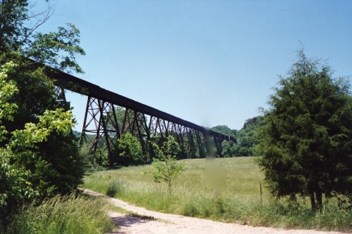 A tall, rusted railway trestle bridge spans a green field under a clear blue sky, surrounded by trees.
