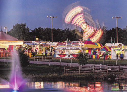A vibrant fair scene at dusk, featuring colorful tents, rides, and a fountain with illuminated water.