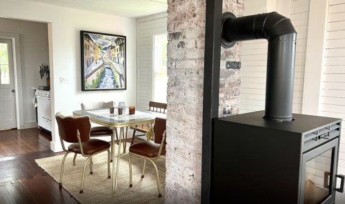 A cozy dining area with a table and chairs, featuring a brick column and a black stove, illuminated by natural light.