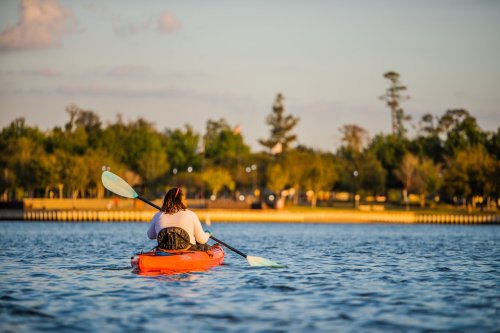 A person paddles a kayak on a calm lake, surrounded by trees and a sunset sky.