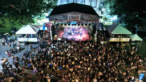 Aerial view of a large crowd enjoying a live band performance at an outdoor festival with tents and stage lights.