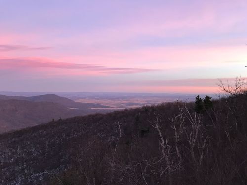 A scenic view of mountains and valleys at sunset, with soft pink and purple hues in the sky.