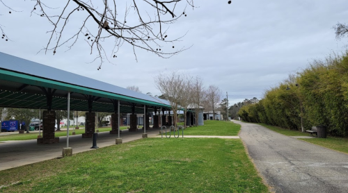 A pathway beside a covered pavilion, with trees and a grassy area under a cloudy sky.