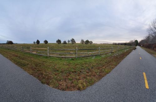 A wide view of a grassy field bordered by a wooden fence, with a paved path running alongside it under a cloudy sky.