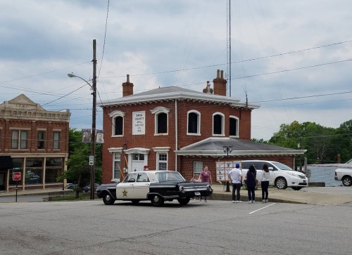 A vintage police car parked in front of a historic brick building, with people gathered nearby on a cloudy day.