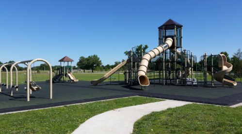 A playground with slides, climbing structures, and swings on a sunny day, surrounded by green grass.