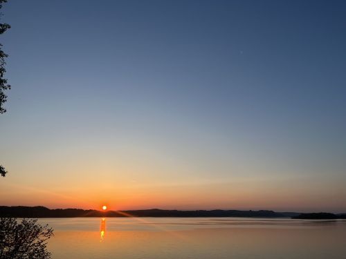 Sunset over a calm lake, with silhouettes of trees and hills against a colorful sky.