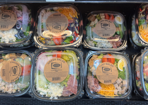 A variety of pre-packaged salads in clear containers, including Greek, Cobb, and Chef salads, displayed in a cooler.