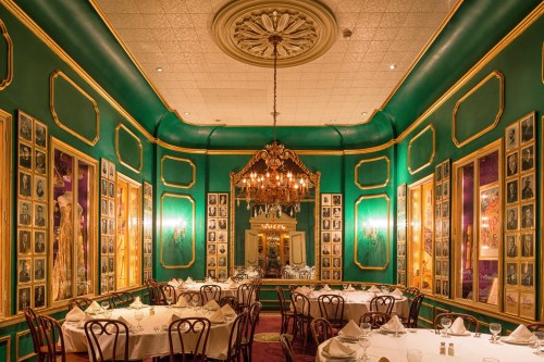 Elegant restaurant interior with green walls, framed photos, chandeliers, and white tablecloths. Cozy and sophisticated ambiance.