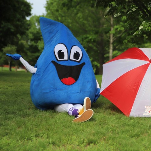 A large blue water droplet mascot sitting on grass next to a red and white umbrella, smiling and waving.