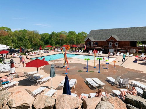A sunny pool area with colorful water features, lounge chairs, and people enjoying the water and surrounding greenery.