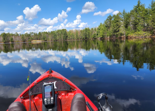 A red kayak on calm water, reflecting clouds and trees, with fishing gear visible in the foreground.