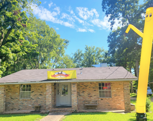A brick building with a bright yellow sign and an inflatable waving figure outside, surrounded by green trees and blue sky.