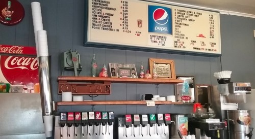 A diner counter with a menu board, soda fountain, and shelves displaying drinks and condiments.