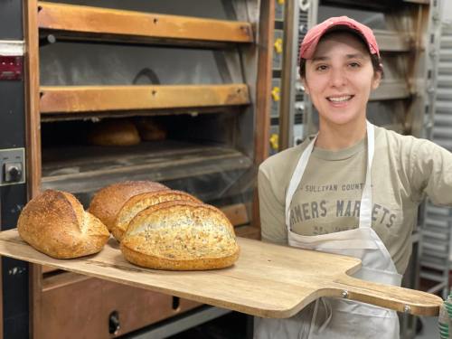 A smiling baker holds a wooden paddle with freshly baked loaves of bread in front of an oven.