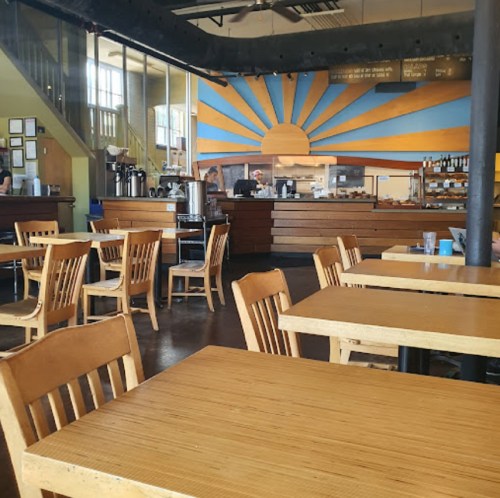 A cozy café interior with wooden tables, chairs, and a sunburst mural on the wall, featuring a counter with baked goods.
