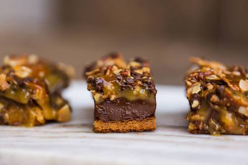 Close-up of layered dessert bars with a chocolate base, caramel, nuts, and a crunchy topping on a marble surface.