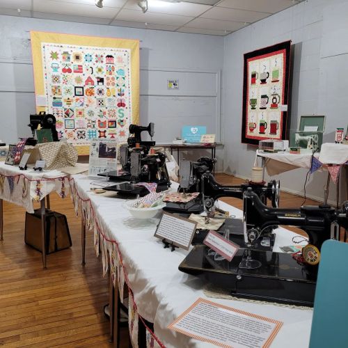 A display of vintage sewing machines and quilts in a museum setting, with tables adorned in fabric and informational signs.