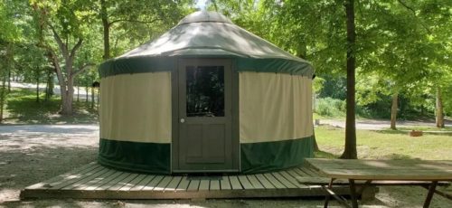 A yurt with a green and beige exterior, set in a wooded area with trees and a gravel path nearby.