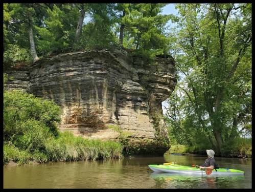 A person in a kayak paddles near a rocky cliff surrounded by lush greenery and trees along a calm river.